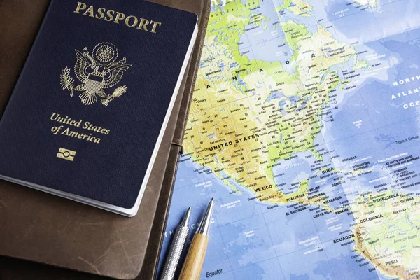 The foil-stamped dark blue front cover of an American passport with leather-covered travel notebooks and writing pen set on a world map background.