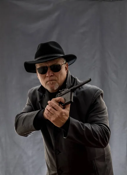 A man in black clothes, a black leather coat, hat. Ganster with a cigar and weapons, a Mauser system pistol. Mafiosi in black glasses on a dark background.  dangerous, unfriendly, evil, rude man. Cowboy felon