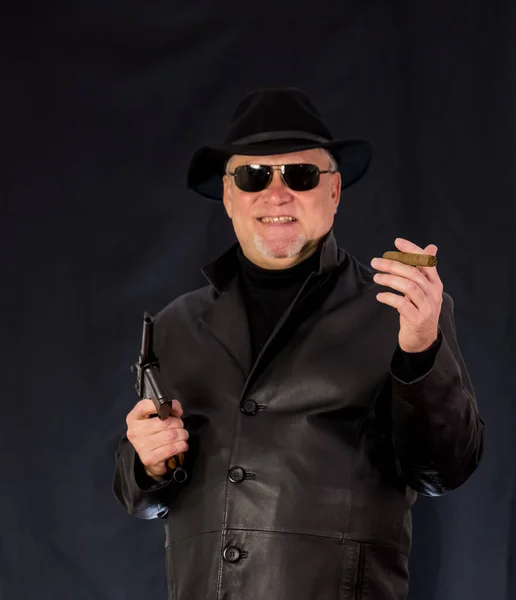 A man in black clothes, a black leather coat, hat. Ganster with a cigar and weapons, a Mauser system pistol. Mafiosi in black glasses on a dark background.  dangerous, unfriendly, evil, rude man. Cowboy felon