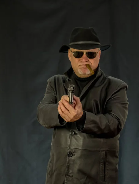 A man in black clothes, a black leather coat, hat. Ganster with a cigar and weapons, a Mauser system pistol. Mafiosi in black glasses on a dark background.  dangerous, unfriendly, evil, rude man. Cowboy felon
