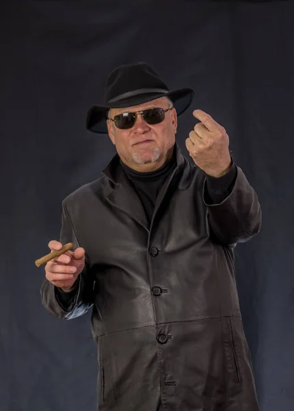 A man in black clothes, a black leather coat, hat. Ganster with a cigar and weapons, a Mauser system pistol. Mafiosi in black glasses on a dark background.  dangerous, unfriendly, evil, rude man. Cowboy felon