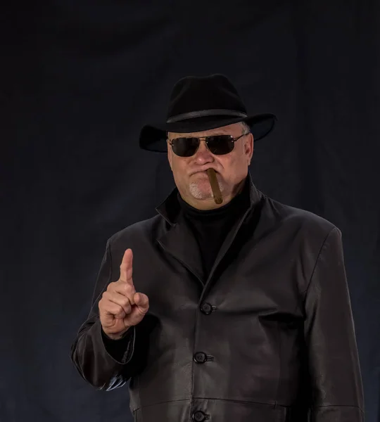 A man in black clothes, a black leather coat, hat. Ganster with a cigar and weapons, a Mauser system pistol. Mafiosi in black glasses on a dark background.  dangerous, unfriendly, evil, rude man. Cowboy felon