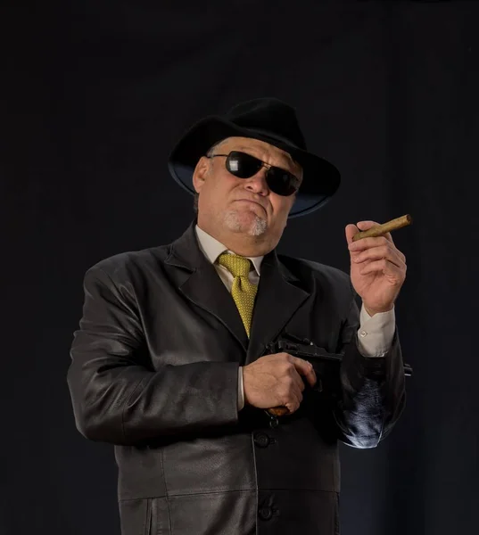 A man in black clothes, a black leather coat, hat. Ganster with a cigar. Mafiosi in black glasses on a dark background. Severe, strong, stylish, impressive, dangerous, unfriendly, evil, rude man
