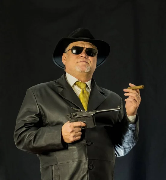 A man in black clothes, a black leather coat, hat. Ganster with a cigar. Mafiosi in black glasses on a dark background. Severe, strong, stylish, impressive, dangerous, unfriendly, evil, rude man