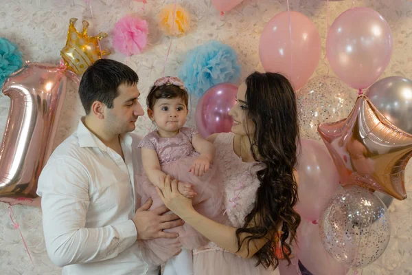 Baby's birthday celebration. The girl was one year old. Happy parents. The child looks away. All are dressed in elegant clothes. Young, beautiful married couple and a child of Caucasian ethnicity. Against the background of balloons, in pink colors.