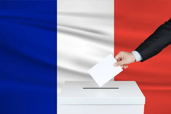 Election in France. The hand of man putting his vote in the ballot box. Waved France flag on background.