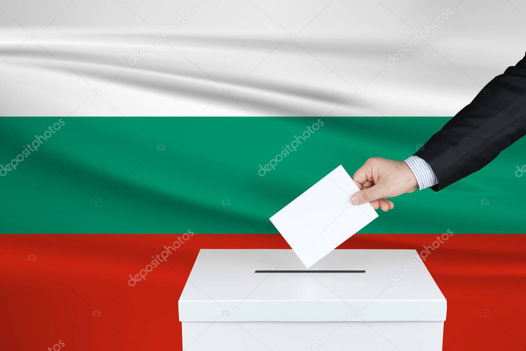 Election in Bulgaria. The hand of man putting his vote in the ballot box. Waved Bulgaria flag on background.