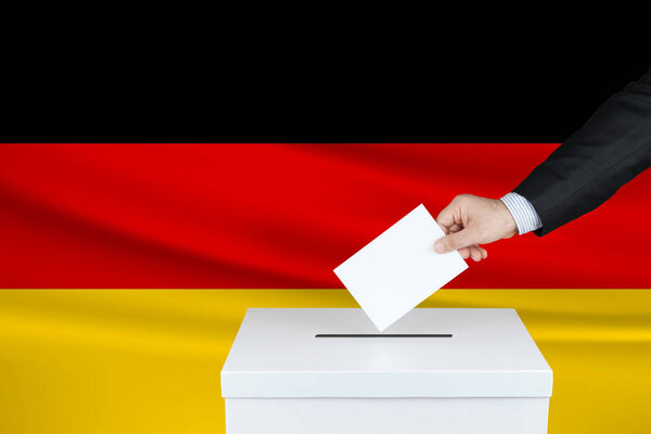 Election in Germany. The hand of man putting his vote in the ballot box. Waved Germany flag on background.                               