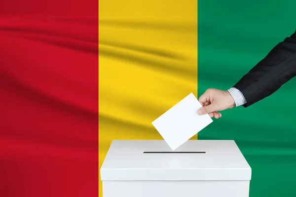 几内亚的选举 把他的选票放进投票箱的人的手 背景上飘扬的几内亚国旗 — 图库照片