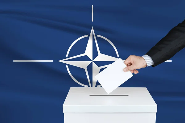 Election in North Atlantic Treaty Organization. The hand of man putting his vote in the ballot box. Waved North Atlantic Treaty Organization flag on background.