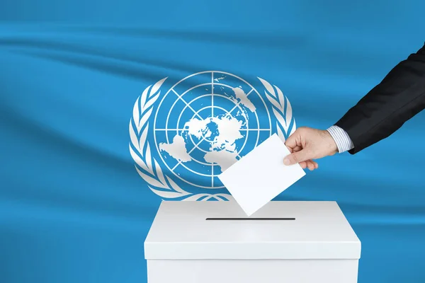 111 联合国的选举 把他的选票放进投票箱的人的手 背景上飘扬的联合国旗帜 — 图库照片