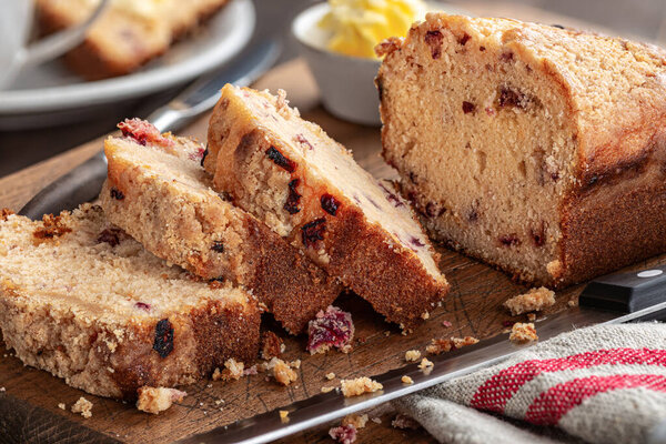 Closeup of sliced cranberry orange loaf cake with a bowl of butter in background on a rustic wooden cutting board