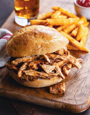 Izgara tavuklu sandviçin yakın plan, arka planda patates kızartması ve ahşap kesim tahtasının üstünde.