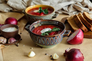 Bej bir zemin üzerinde borş çorbası. Kil tabakta sarımsak, soğan, biber, ekşi krema ve ekmek.