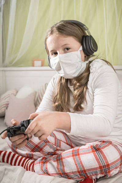 Girl in a mask playing on a game console during quarantine on the bed.