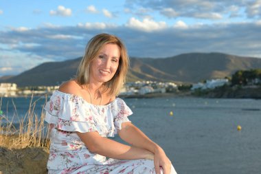 Happy woman sitting in front of a coastal landscape of the northern mediterranean on a summer afternoon with wind looking at the camera smiling with copy space