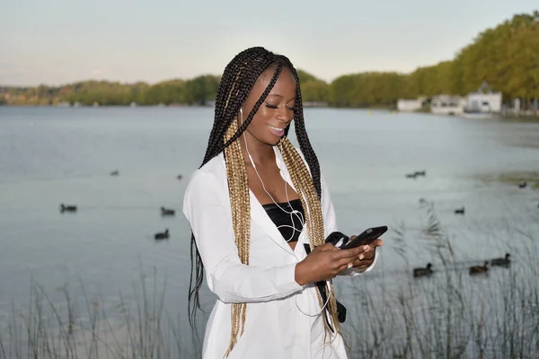 Afro-schwarzes Mädchen mit langen schwarzen und blonden Zöpfen, das ihr Telefon mit Kopfhörern und einem See im Hintergrund benutzt — Stockfoto