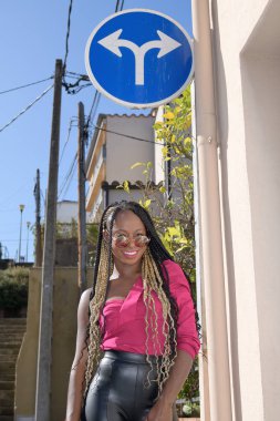 Küçük bir kasabada trafik levhasının yanındaki kameraya bakarken uzun siyah ve sarı örgülü ve güneş gözlüklü mutlu afro-stil siyah kız gülümsüyor.