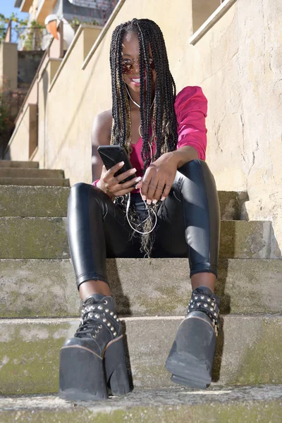 Glückliches schwarzes Mädchen im Afro-Stil mit langen schwarzen und blonden Zöpfen und Sonnenbrille sitzt auf der Treppe und lächelt in die Kamera, während sie ihr Telefon und Kopfhörer benutzt — Stockfoto