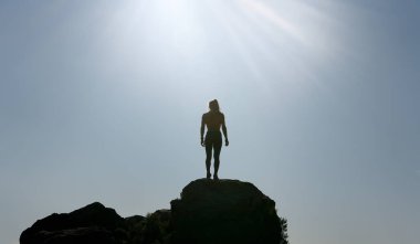 Sporcu kadın güneşin altında, dağın en yüksek kayasında, arka planda gökyüzü ile dinleniyor.