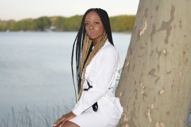 Afro kız kameraya uzun siyah ve sarı örgülü ve arka planda gölü olan pozlar veriyor.