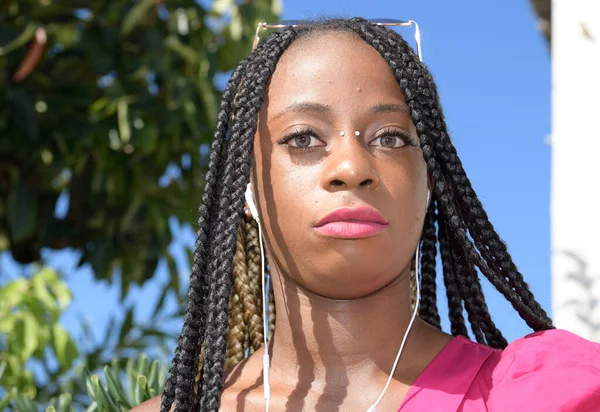 Porträt eines schwarzen Mädchens im Afro-Stil mit langen schwarzen und blonden Zöpfen. Sie blickt mit ängstlichem Gesichtsausdruck in die Kamera. sie benutzt Kopfhörer und Sonnenbrille — Stockfoto