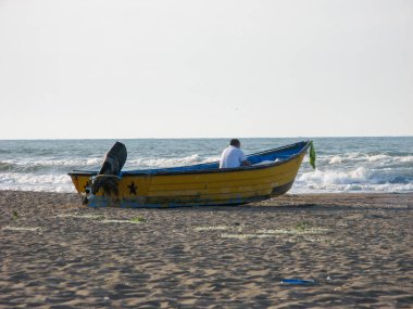 Eski sarı ahşap balıkçı teknesi ve balıkçı İran 'da, deniz kenarı kasarının yakınında kıyıda karaya oturmuş. Deniz kıyısındaki bir teknede oturan bir adam..