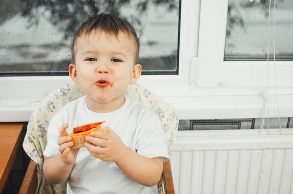 baby in the kitchen in a white shirt eating a sandwich with sausage