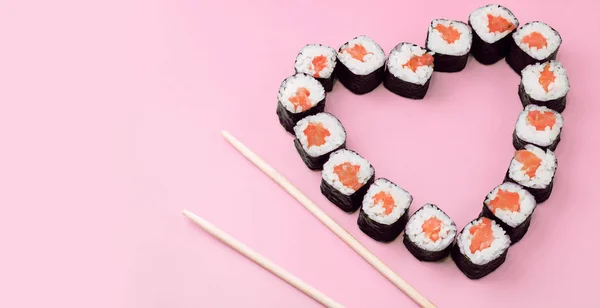 Salmon sushi rolls laid out in the shape of a heart on a pink background. The concept of Japanese cuisine for Valentine\'s Day, greeting card, banner. Copy space