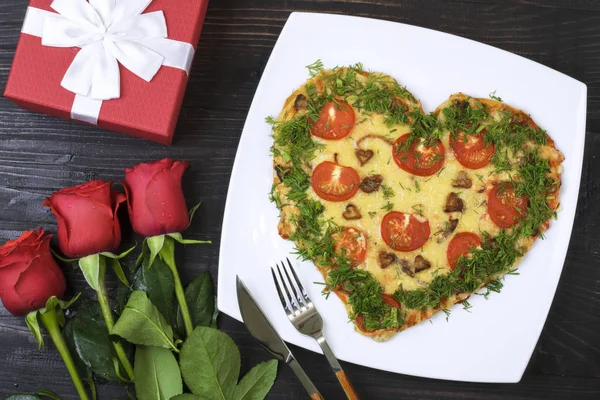 Valentine\'s day food. Heart shaped pizza next to a gift and red roses, on a dark wooden background. Valentine\'s day concept Top view. Copy space