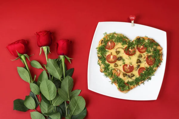 Valentine's day food. Heart shaped pizza next to a red roses, on a red background. Valentine's day concept. Top view. Copy space