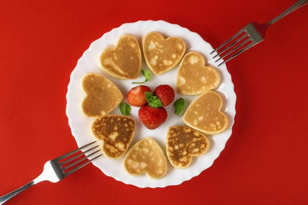 Creative breakfast for Valentine\'s Day. Heart shaped pancakes with strawberries on a plate with forks, on a red background. Valentine\'s day background. Top view. Flat lay. Copy space