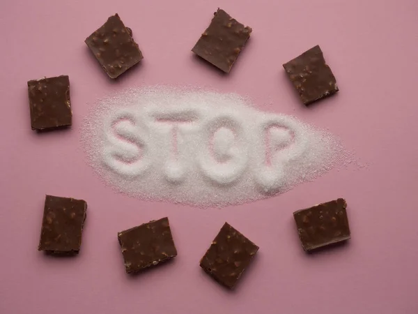 Heap of white sugar with word STOP hand written on it with pieces of chocolate on a pink background top view. Stop diabetes and excessive sugar intake concept. Healthcare, unhealthy diet concept.