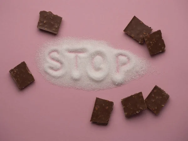 Heap of white sugar with word STOP hand written on it with pieces of chocolate on a pink background top view. Stop diabetes and excessive sugar intake concept. Healthcare, unhealthy diet concept.