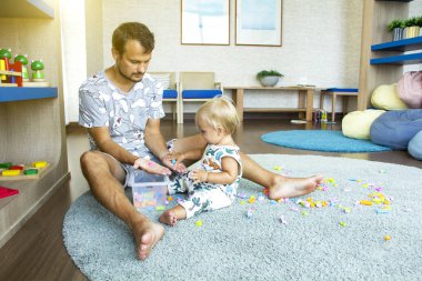 Babam anaokulunda tatlı sarışın bebeğiyle büyük gri halıda tuğlalarla oynuyor..