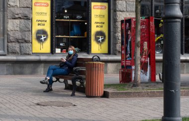 Kiev, Ukrayna, 27 Mart 2020, yüz koruyucu maskeli genç Ukraynalı kadın cep telefonuyla bankta oturuyor.