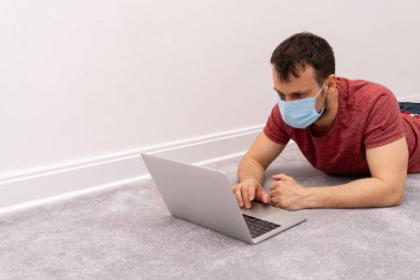 Yüz koruma maskeli bir adam yerde yatar ve laptopta çalışır.
