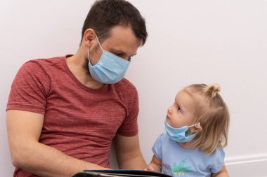 Tıbbi maskeli kız, maskeli ve kitap okuyan babaya bakar.