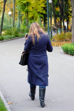 Koyu mavi paltolu ve siyah çizmeli bir kadın bükülmüş kolunda bir çantayla park yolunda yürüyor. Sonbahar parkının arka sokağı, bir iş kadını. Sonbaharda şehir parkı.