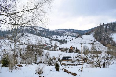 Gri gökyüzüne karşı karla kaplı dağlar. Dağlardaki bir köyde kış manzarası. Tepelerde birkaç ahşap ev var..
