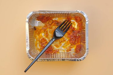 Yemek artıklarıyla birlikte alüminyum folyo kabı ve siyah plastik çatal. Tek kullanımlık alüminyum yiyecek kabı üst görünümü. Kullanılan kirli tek kullanımlık sofra takımı.