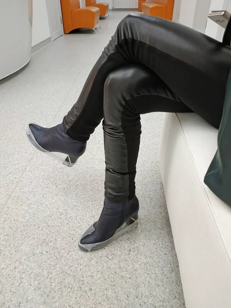 Legs of a girl in black boots and disposable shoe covers. Close-up leather pants and high heel boots in shoe covers. Girl sitting waiting for a doctor\'s appointment at the clinic.