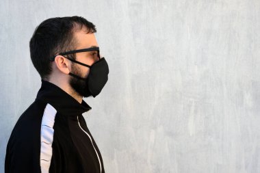 Siyah maskeli ve gözlüklü bir adam gri bir duvarın karşısında kameraya yan dönmüş duruyor. Salgının teması ve virüsün tacı enfeksiyonlara karşı korunmak için maske takmak. Model