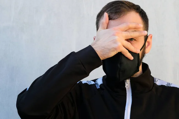 A guy in a black cotton face mask. A man is standing in a black sports jacket and has covered his face with his hand. Pandemic corona virus precautions against infection.