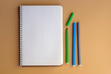 Beyaz not defteri ve renkli keçeli kalemler üst görünümde. Bej arka planda boş bir defter sayfası. İlham ara, liste yap, notlar veya çizimler için bir yay üzerine not al.