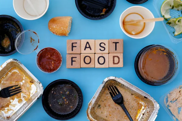 On a blue background are empty aluminum containers and plastic utensils. The inscription fast food top view. Food delivery, empty dirty dishes from under the meal. Harm from fast food and obesity.