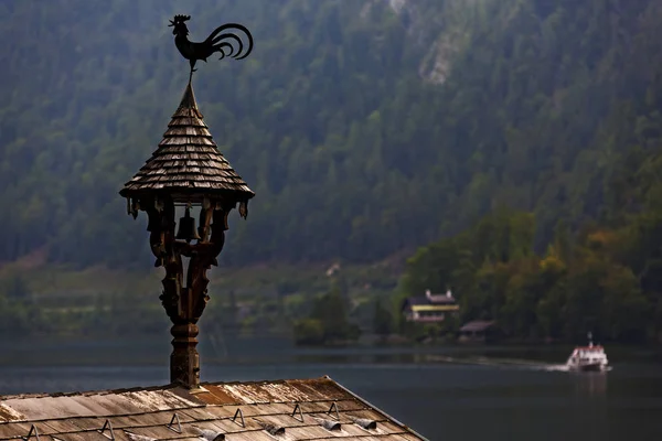 Paleta vieja en forma de gallo en una pequeña torre de madera en el techo de la casa temprano en la mañana contra los Alpes y el lago — Foto de Stock