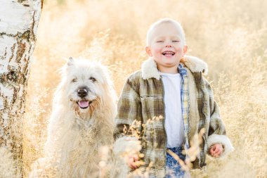Mutlu gülümseyen sarışın çocuk çiftlikte köpek arkadaşıyla yürüyor.