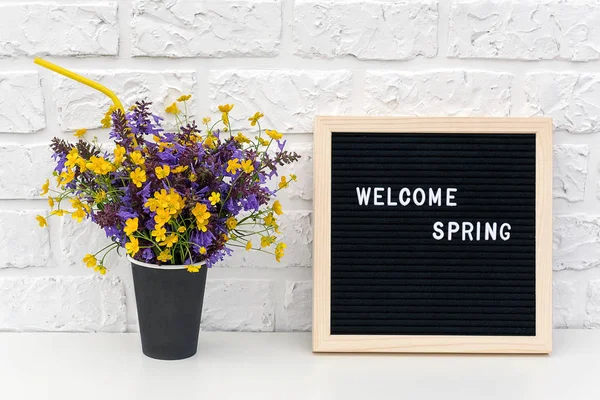 Text Hello Spring on black letter board and bouquet of colored flowers in black paper coffee cup with cocktail straw on background white brick wall. Concept Springtime mood — Stock Photo, Image