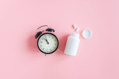 İki hap, beyaz şişe ve pembe arka planda siyah alarm saati. Insomnia kavramı, uyku sorunları, ilaç alıp tedavi olma zamanı. Üst Görünüm Düz yatay Boşluğu Kopyala